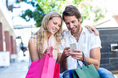 Smiling couple with shopping bags sitting and using smartphone