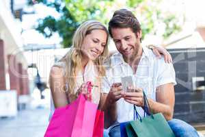 Smiling couple with shopping bags sitting and using smartphone