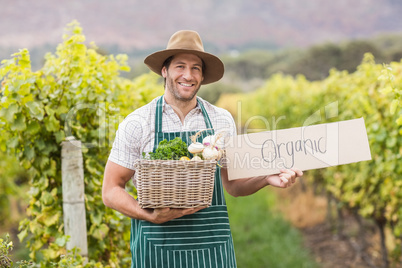 Young happy farmer holding a basket of vegetables and a sign