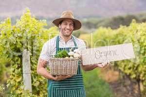 Young happy farmer holding a basket of vegetables and a sign