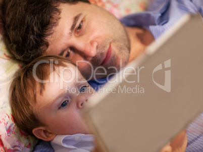 Man and little boy playing with tablet in bed