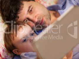 Man and little boy playing with tablet in bed