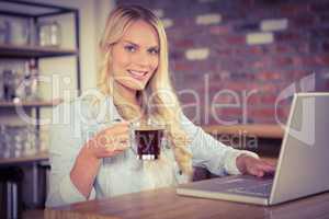 Smiling blonde drinking coffee and using laptop