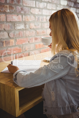 Smiling blonde drinking coffee and writing on sheet of paper