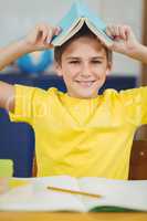 Smiling pupil holding book on head in a classroom