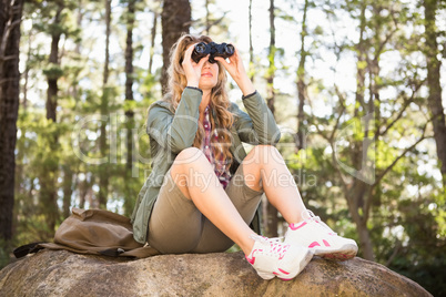 Blonde hiker looking through binoculars and sitting on stone