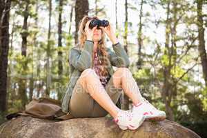 Blonde hiker looking through binoculars and sitting on stone