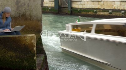 Woman using laptop by the canal with sailing boat