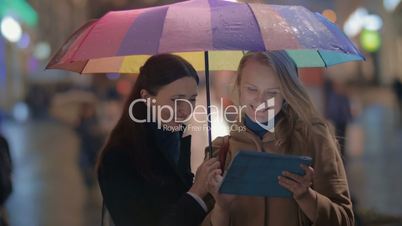 Female friends using touch pad on rainy evening in city