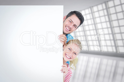 Composite image of smiling young couple hiding behind a blank si