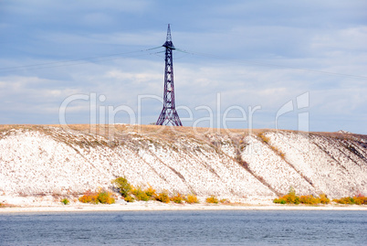 High voltage line and electricity pylon on coastline of river.