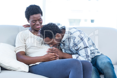 Happy pregnant woman with husband kissing her belly at home