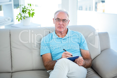 Thoughtful senior man in eyeglasses sitting on sofa with clipboa