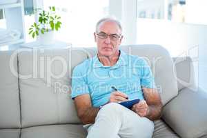 Thoughtful senior man in eyeglasses sitting on sofa with clipboa