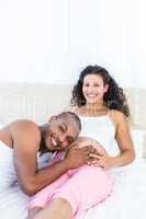 Portrait of man listening to belly of happy pregnant wife on bed