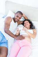 Pregnant wife holding smartphone with husband lying on bed
