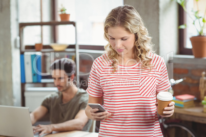 Businesswoman using smartphone while holding coffee in office