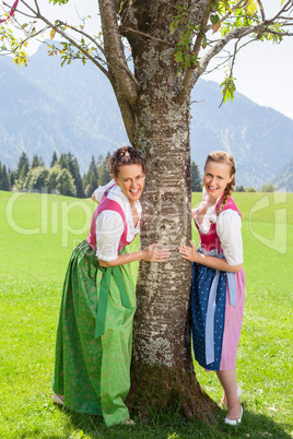 Zwei lächelnde Frauen Im Dirndl halten sich an einem Baum fest.