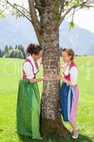 Zwei  bayerische Frauen halten sich an einem Baum fest und lächeln sich an