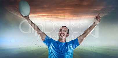 Composite image of happy rugby player in blue jersey holding ball with arms raised