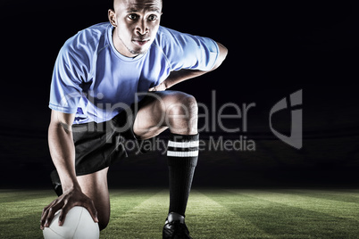 Composite image of serious rugby player kneeling while holding b