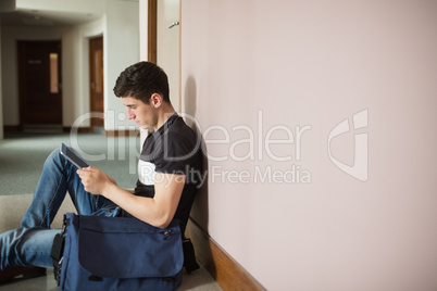 Male student with digital tablet sitting by wall