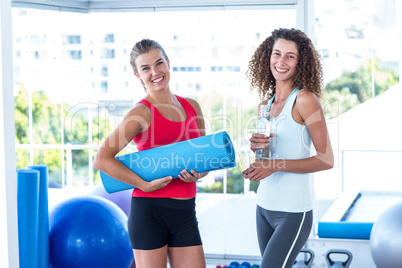 Fit women holding exercise mat and water bottle in fitness studi