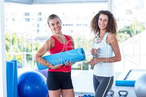Fit women holding exercise mat and water bottle in fitness studi