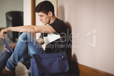 Male student with digital tablet sitting by wall in college