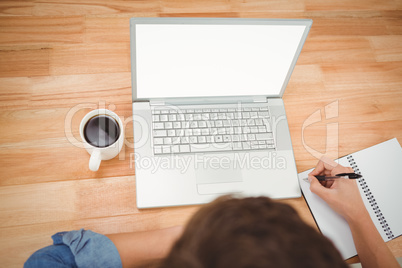 Hipster writing on spiral notebook by laptop at desk in office