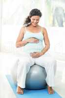 Smiling pregnant woman touching her belly while sitting on exerc