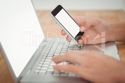 Man holding mobile phone while typing on laptop at desk