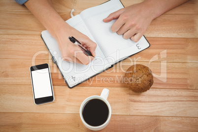 Hipster writing on diary with muffin and coffee at desk