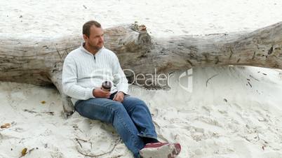 a man sitting in the sand on a fallen tree and drinks a coffee