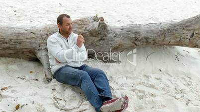 a man sitting in the sand on a fallen tree and eats an apple