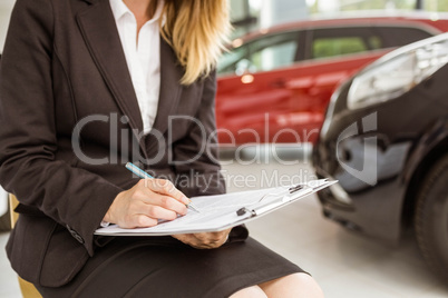 Saleswoman sitting near cars while writing on clipboard