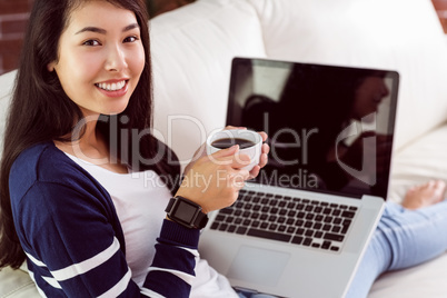 Asian woman relaxing on couch with coffee using laptop