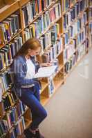 Student reading book in library leaning against bookshelves