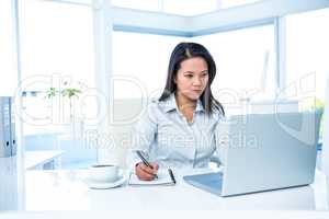 Serious businesswoman writing on notepad and using laptop