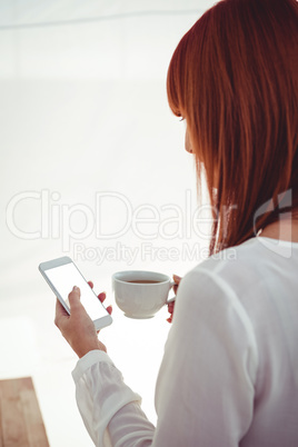 Woman with red hair using smartphone and holding coffee cup