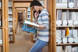 Focused student leaning against bookshelves and reading a book i