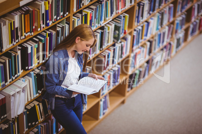 Student reading book in library leaning against bookshelves