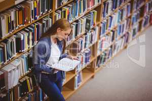 Student reading book in library leaning against bookshelves