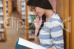 Focused student leaning against bookshelves and reading a book i