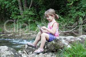 little girl sitting next to a stream and play with tablet