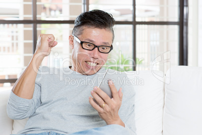 Mature Asian man celebrates success while using smartphone