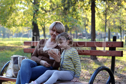 mother and daughter sitting in park and play with tablet