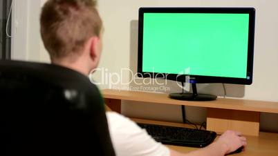 Young man writes on the keyboard and looks at the monitor on the table in room - green screen