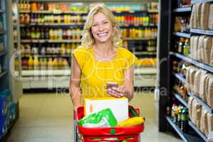 Smiling woman with smartphone pushing trolley in aisle