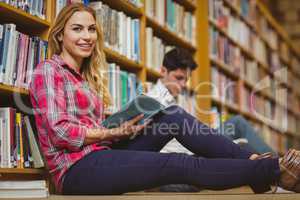 Smiling classmates reading book while leaning on bookshelves